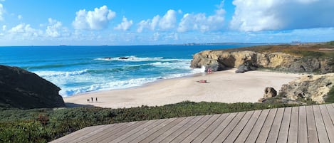 A wooden deck overlooking a beach with waves, a cliff, and a clear sky.