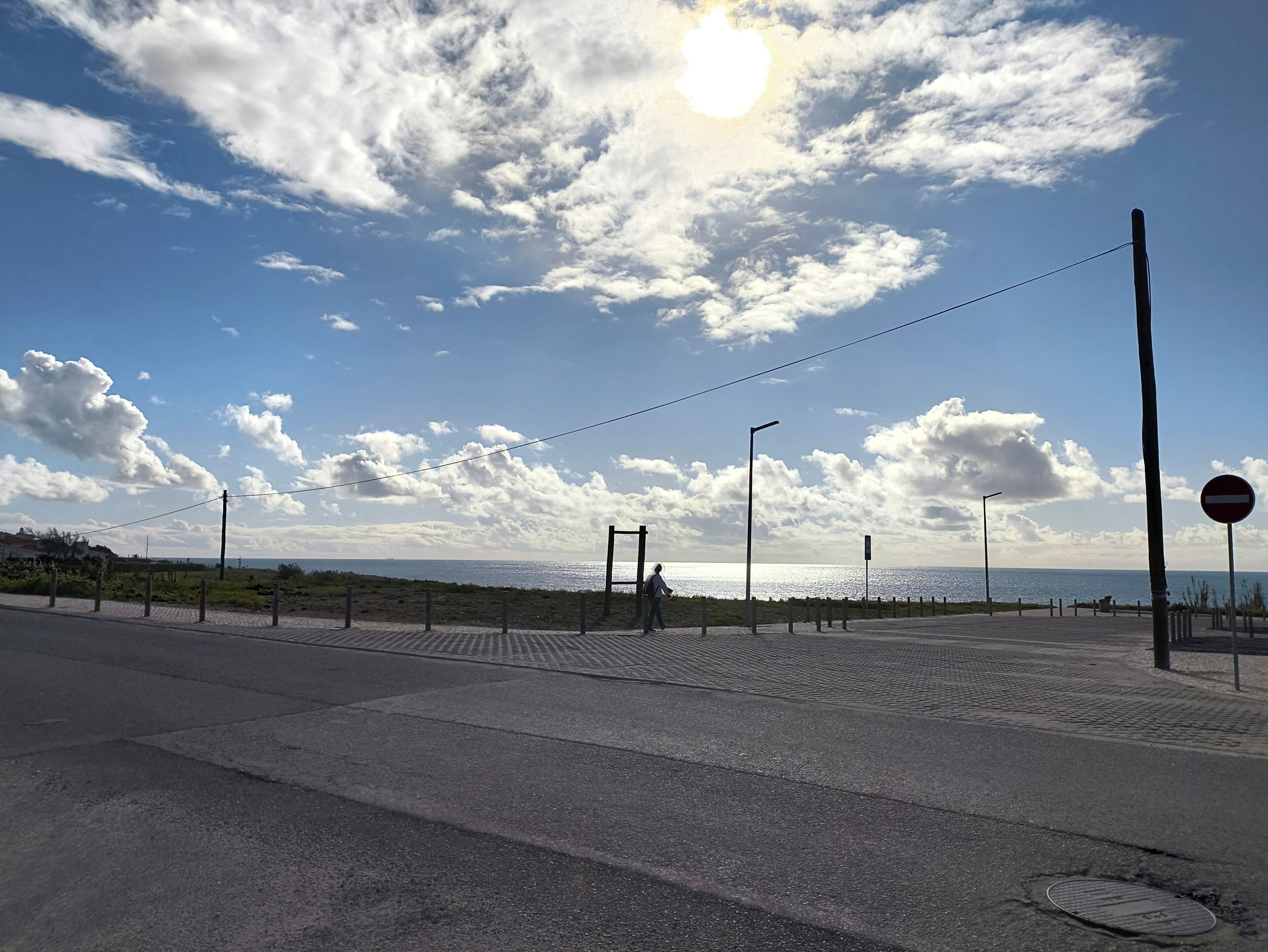Ein gepflasterter Weg mit blauem Himmel und Wolken, ein Strand in der Ferne und ein Verbotsschild.
