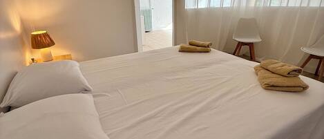 Un dormitorio con cama, almohadas, lámpara, silla y ventana con cortinas.