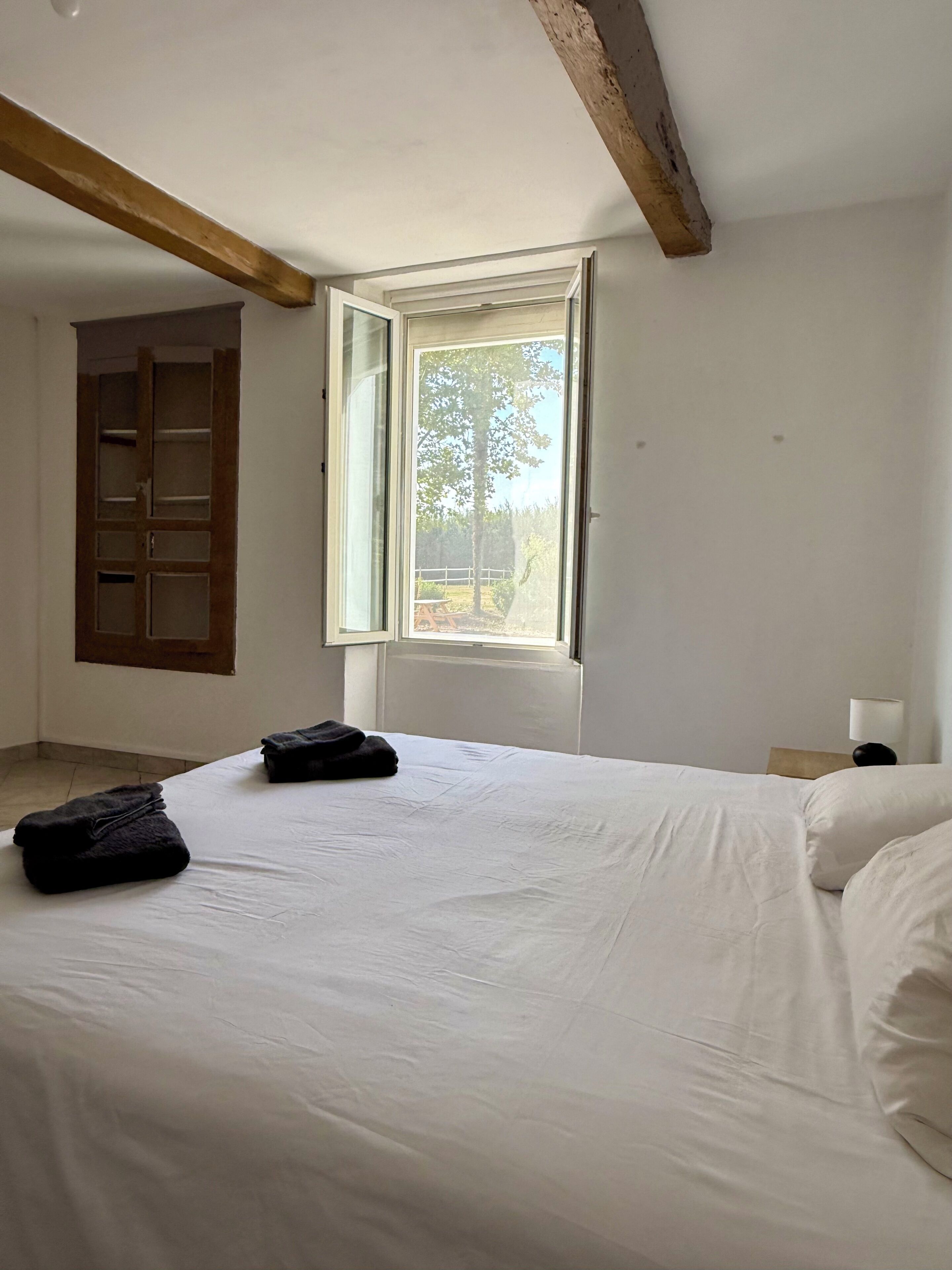 A bedroom with a large bed, a window with an open shutter, a wooden door, and a nightstand with a lamp.