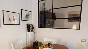 Zona de menjador amb taula de fusta, cadires blanques, un bol de fruita i quadres a la paret.