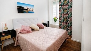 Un dormitori amb un llit, una tauleta de nit, una làmpada, una finestra amb cortines i un quadre penjat a la paret.