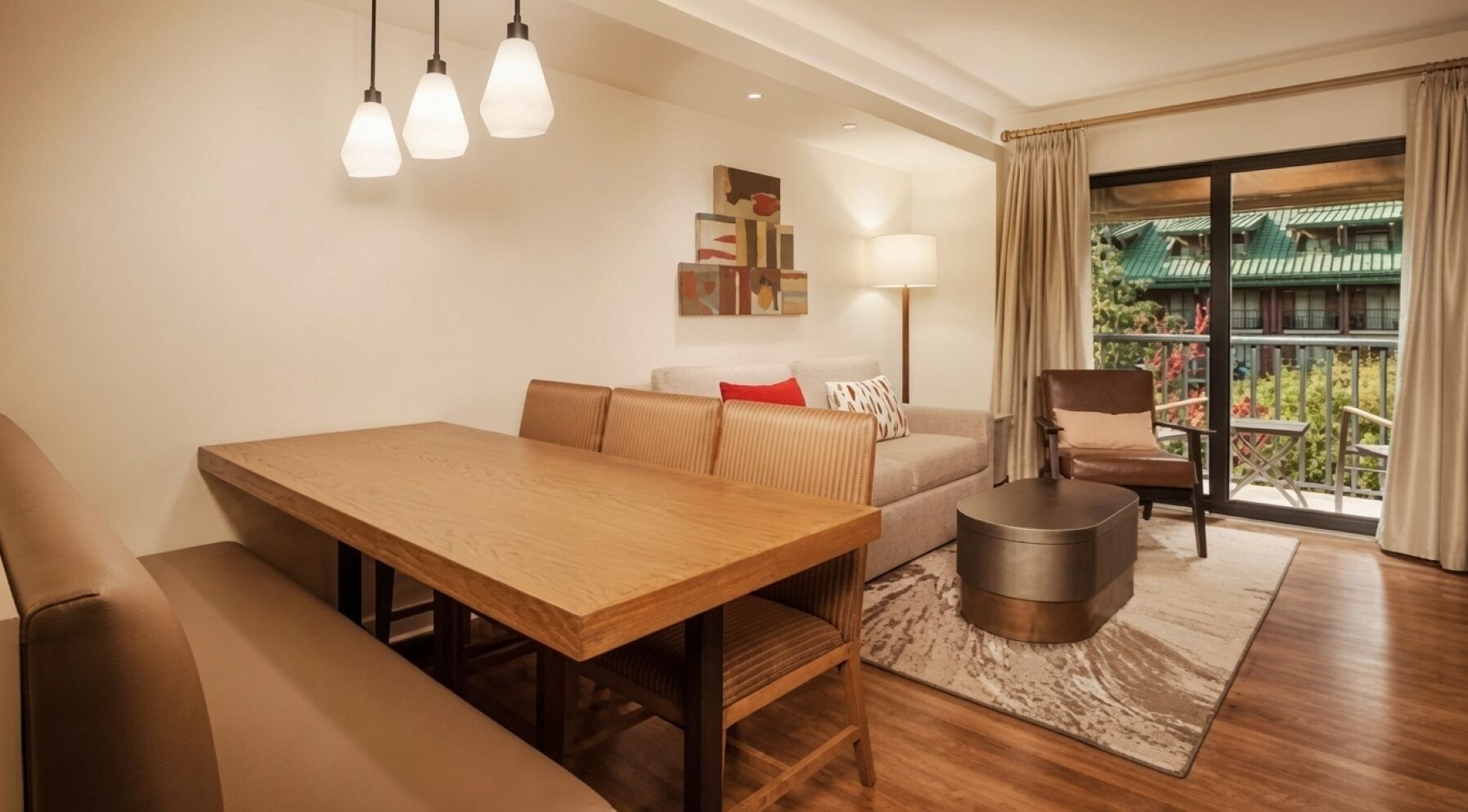Uma sala de estar moderna com uma mesa de jantar de madeira, um sofá, uma cadeira e uma pequena mesa redonda. Há uma varanda com vista para o verde.