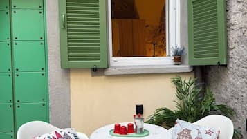 Una piccola area esterna con un tavolo e due sedie, una finestra con persiane verdi e un vaso con una pianta.