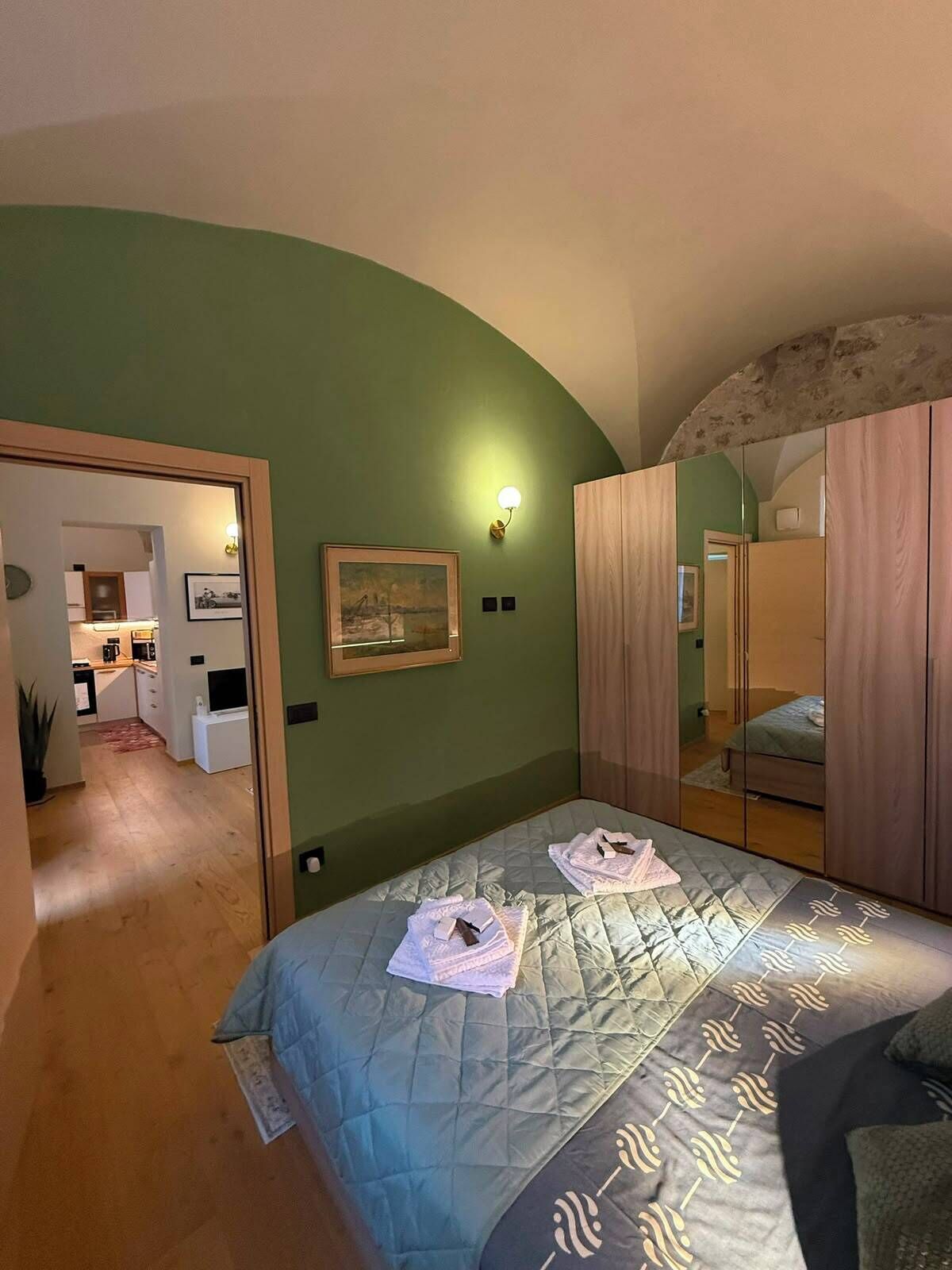 A bedroom with a bed, a mirror, a painting, and a kitchenette in the background.