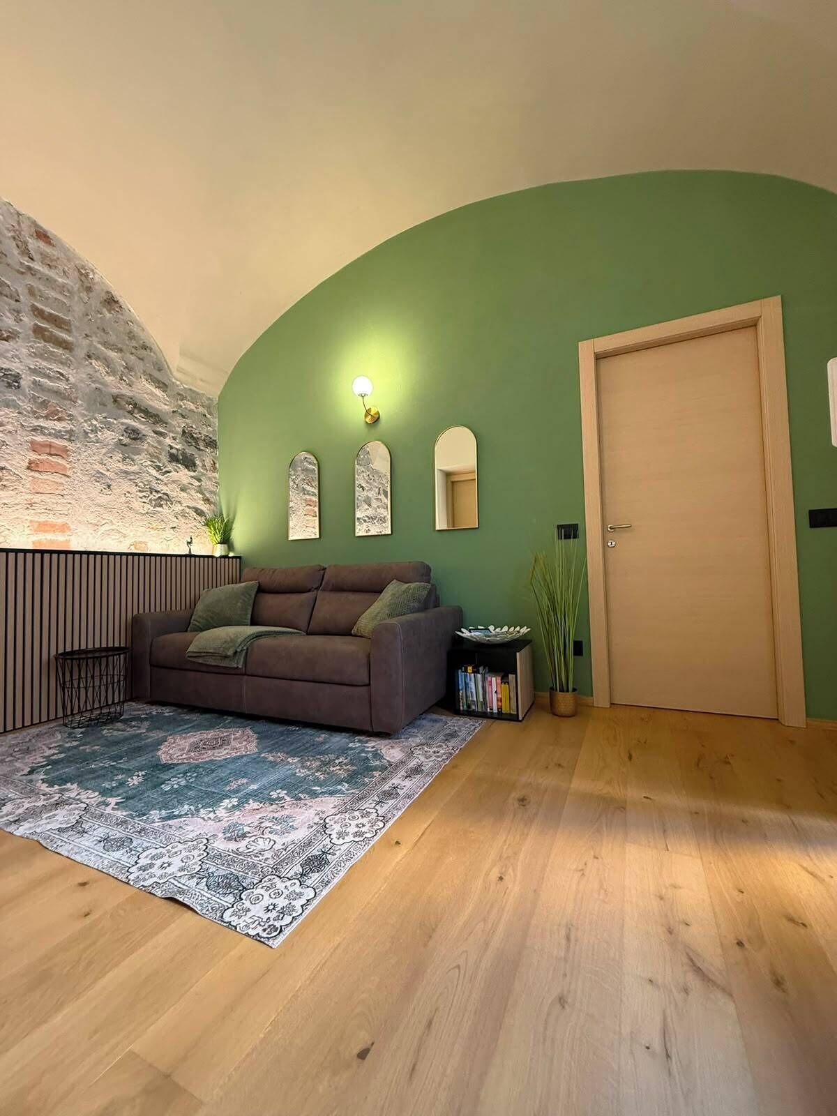 Ein Wohnzimmer mit braunem Sofa, einem gemusterten Teppich, einer grünen Wand, einer Holztür und einer Backsteinwand.