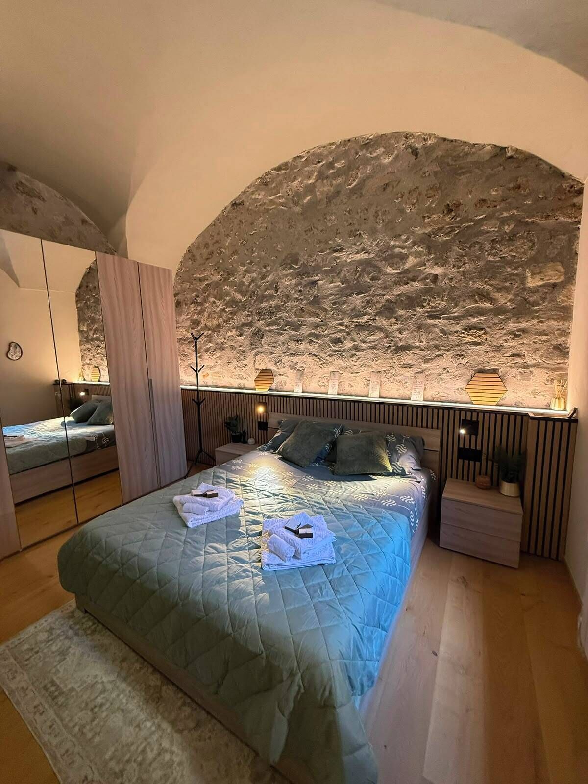 A bedroom with a large bed, a stone wall, a mirror, and bedside tables.
