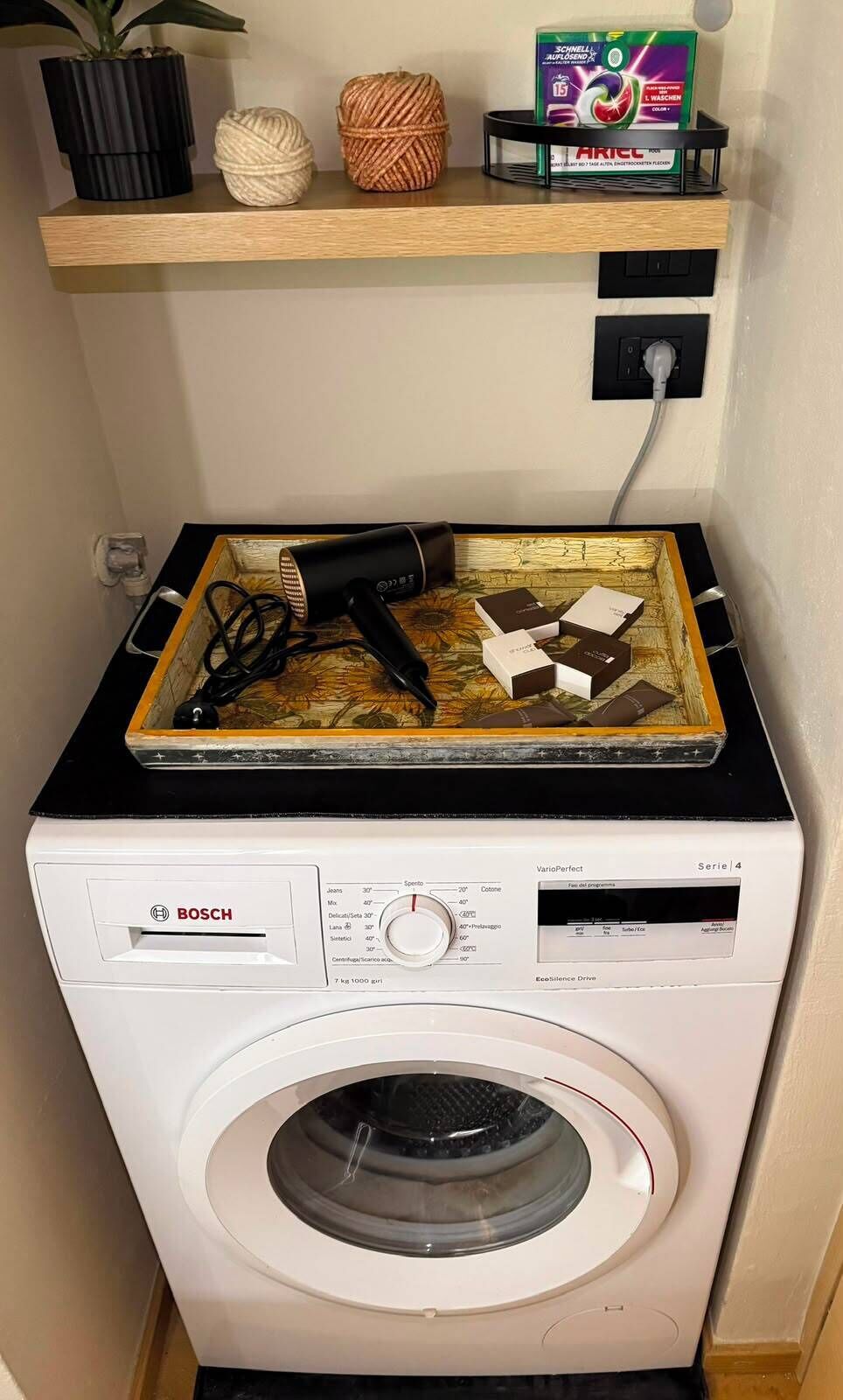 Un lave-linge Bosch avec un étendoir placé sur le dessus, une prise de courant et une étagère sur laquelle se trouvent une plante, une pelote de laine et un paquet de lessive.