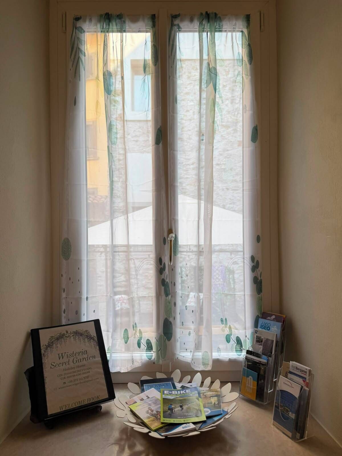 Une fenêtre ornée de rideaux légers à motifs de feuilles, un livre d’accueil et des brochures disposées sur une table.