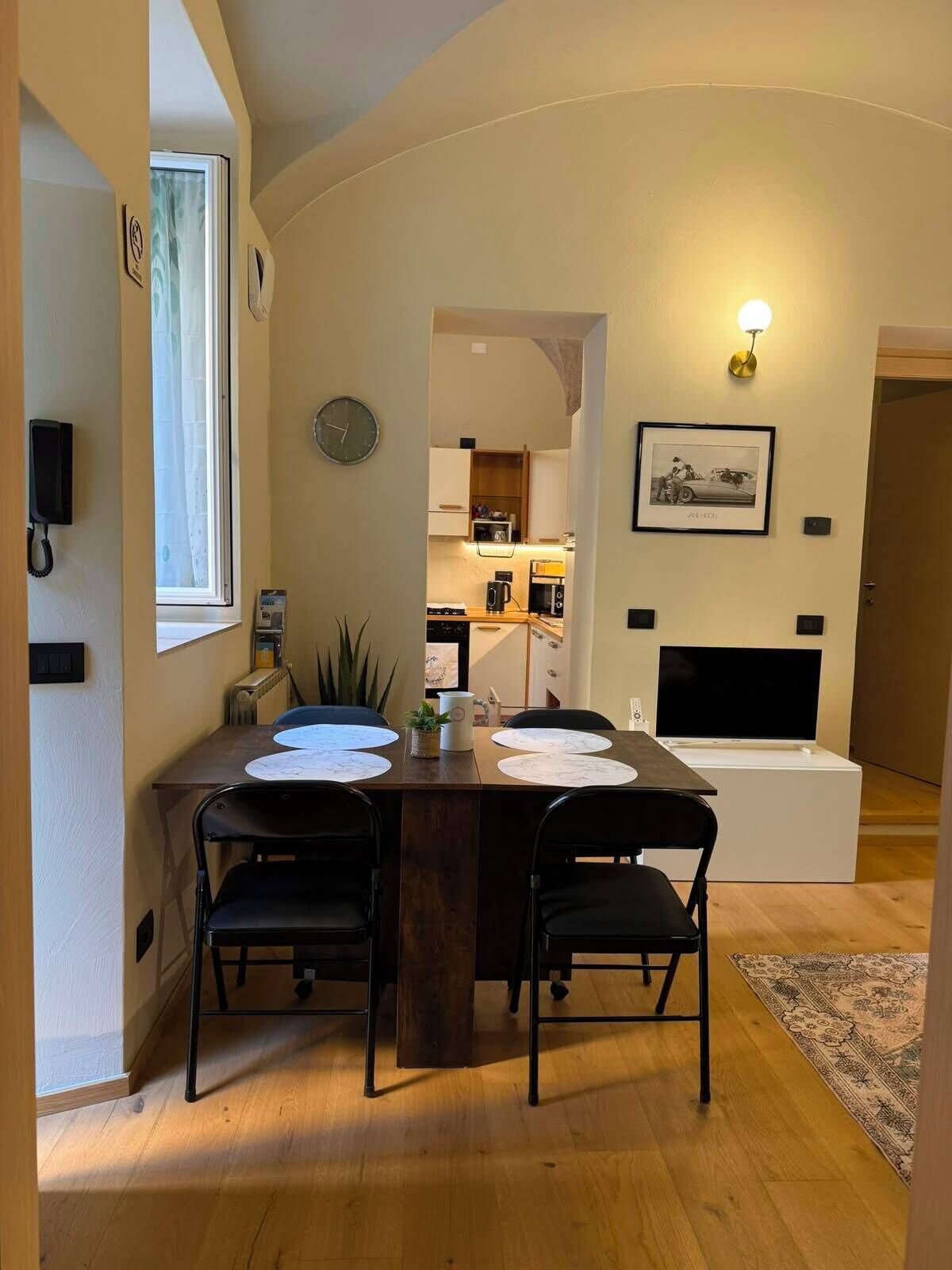 A dining area with a wooden table, four chairs, a clock, and a framed picture on the wall.