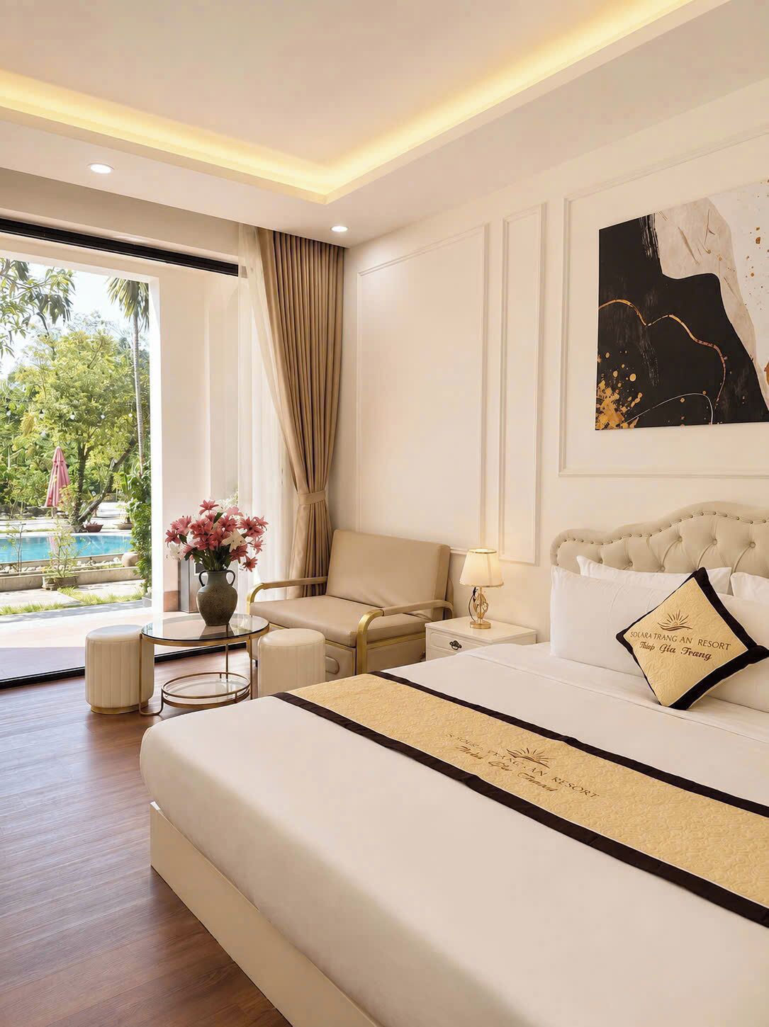 Habitación de hotel con una cama grande, una mesita de noche, una silla, una pequeña mesa con un jarrón de flores y vistas al exterior.