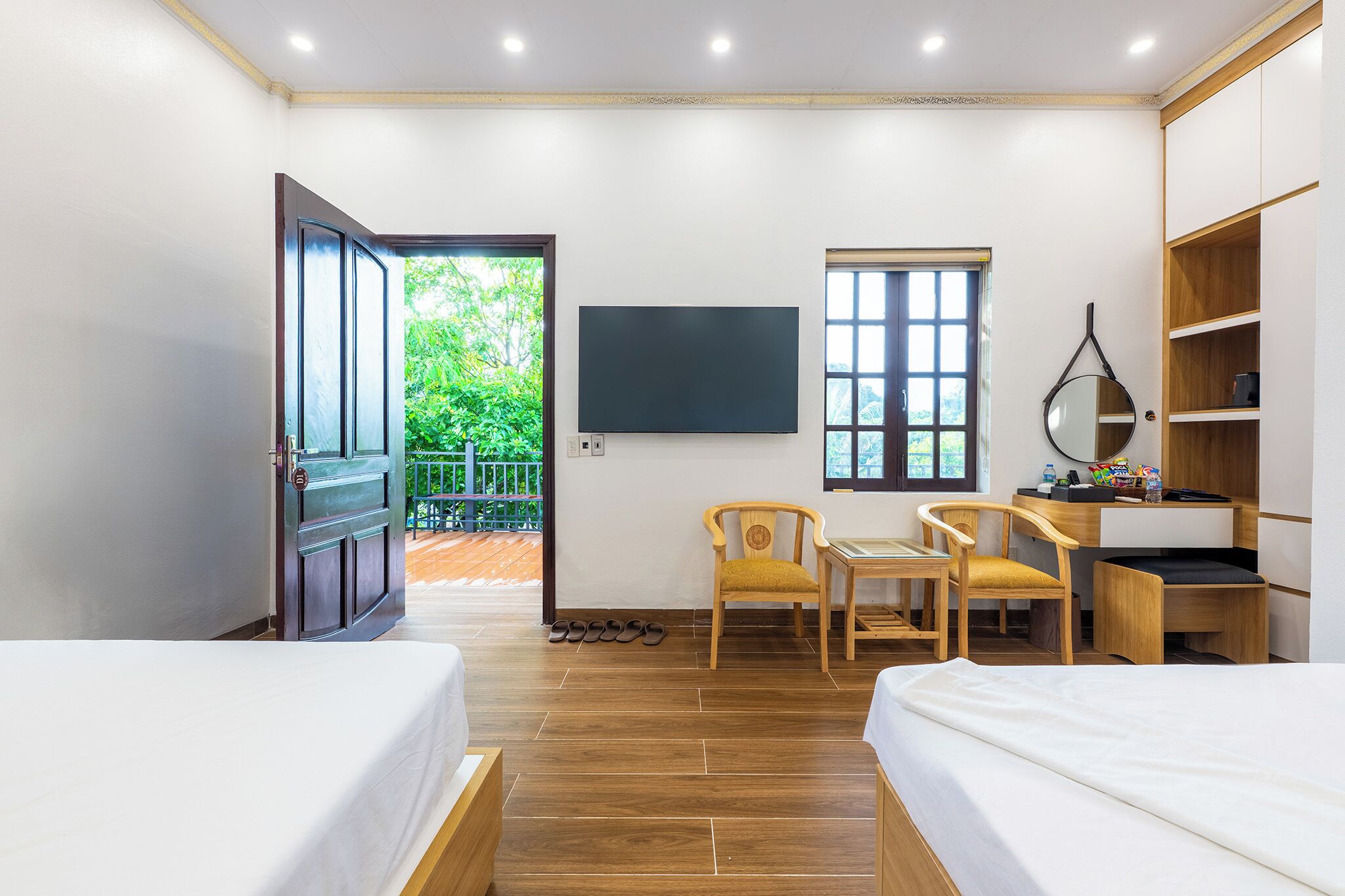 A hotel room with two beds, a desk with a chair, a TV, and a window with a view of greenery.