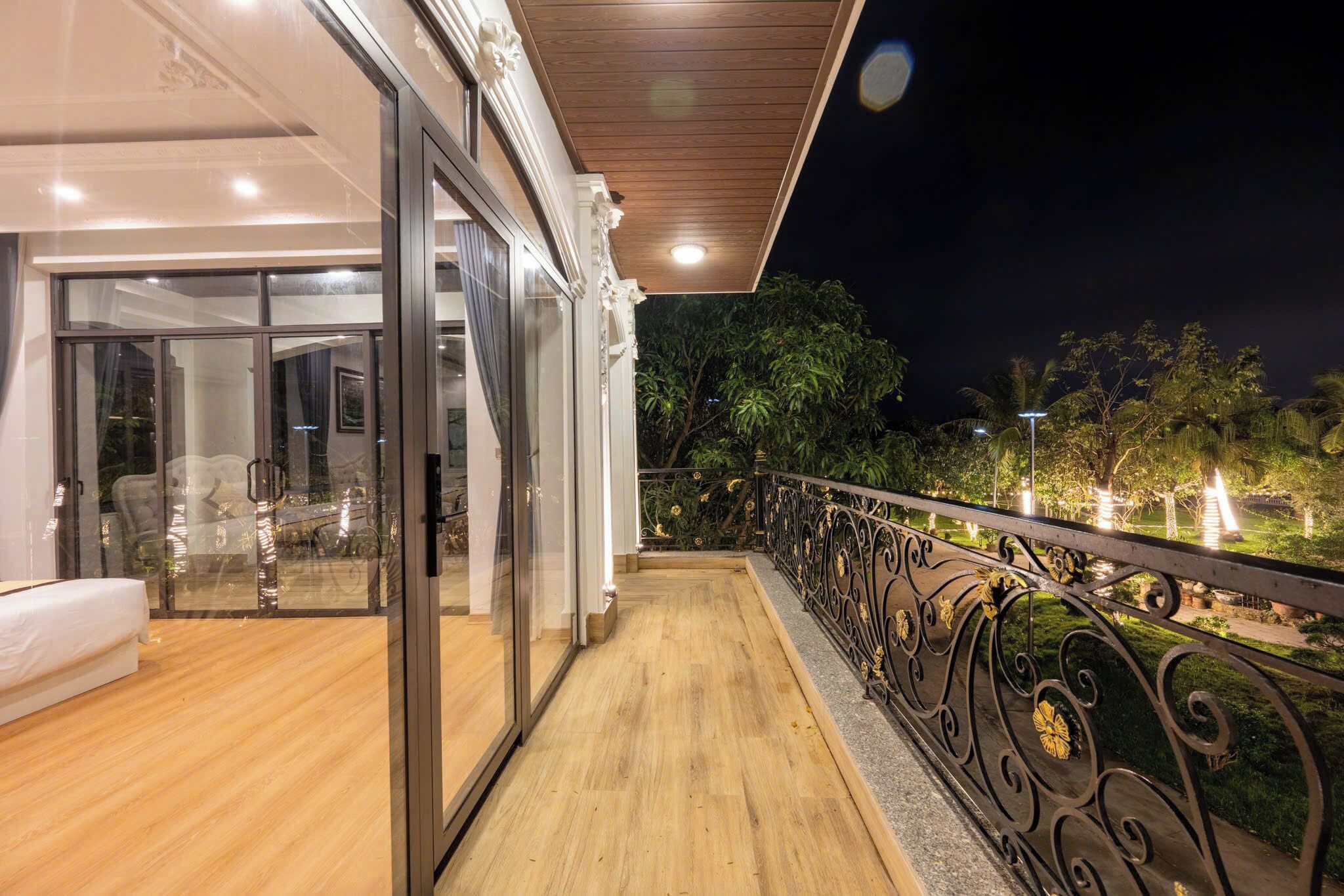 A balcony with a wooden floor and a metal railing, overlooking a garden with trees and lights.