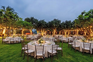 An outdoor dining area with round tables and chairs, surrounded by trees and string lights.