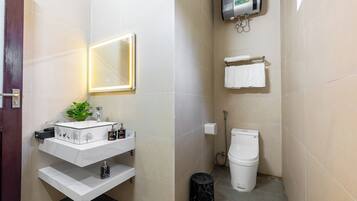 Casa de banho compacta com uma sanita, lavatório branco, espelho e um pequeno contentor de lixo.