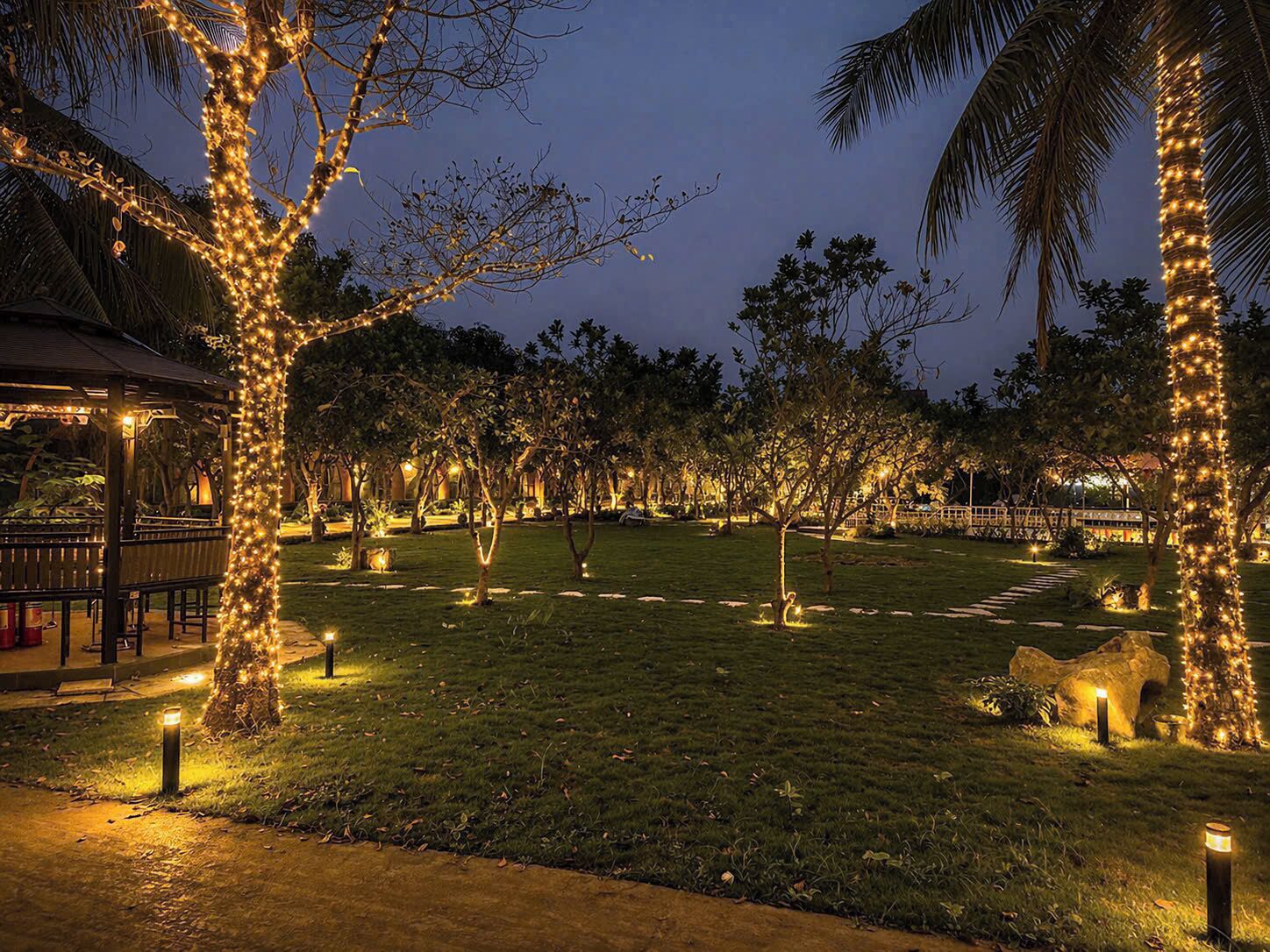 A nighttime outdoor area with trees wrapped in lights, a gazebo, and a grassy lawn.