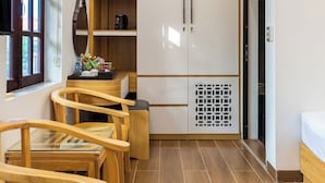 Una habitación compacta con piso de madera, una kitchenette con armarios blancos, un pequeño rincón de descanso con sillas y una ventana con persianas.