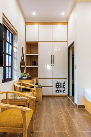 A compact room with a wooden floor, a kitchenette with white cabinets, a small seating area with chairs, and a window with blinds.