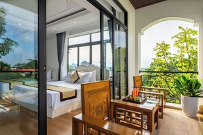 A bedroom with a large window, a balcony with a view of greenery, and a dining area with a table and chairs.