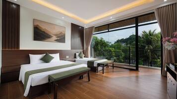 A hotel room with a large bed, a smaller bed, a bench, a desk, and a balcony with a view of greenery.