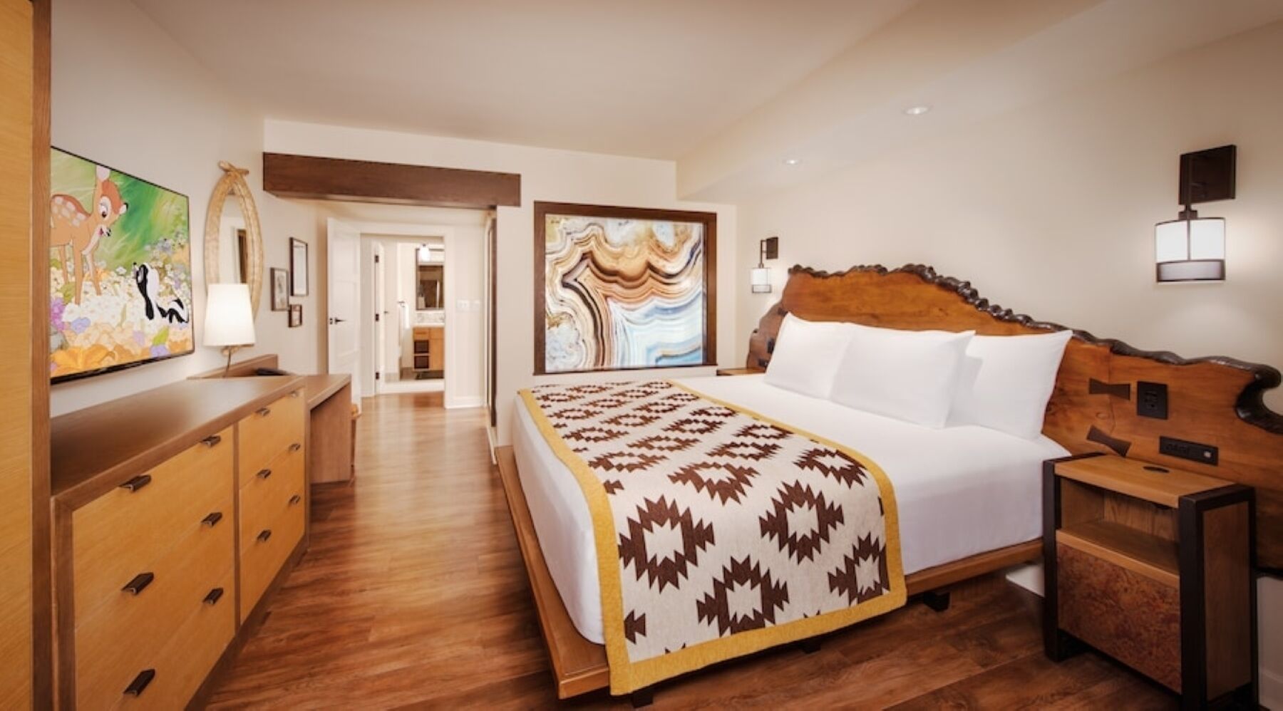 A hotel room with a bed, wooden furniture, a painting, and a view into the hallway.