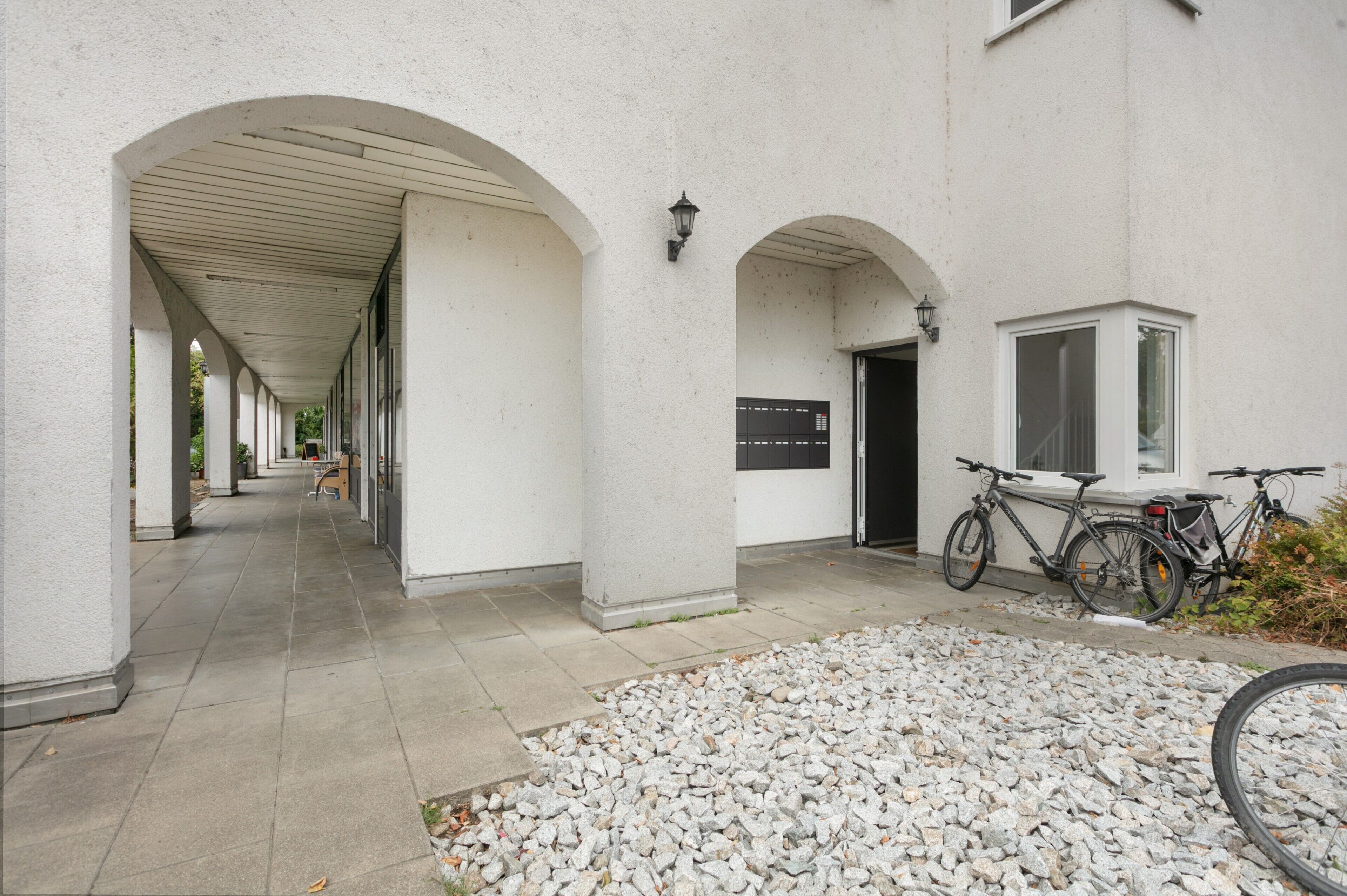 Une allée couverte, dont le sol est en pierre, mène à l’entrée d’un bâtiment devant lequel est garé un vélo.