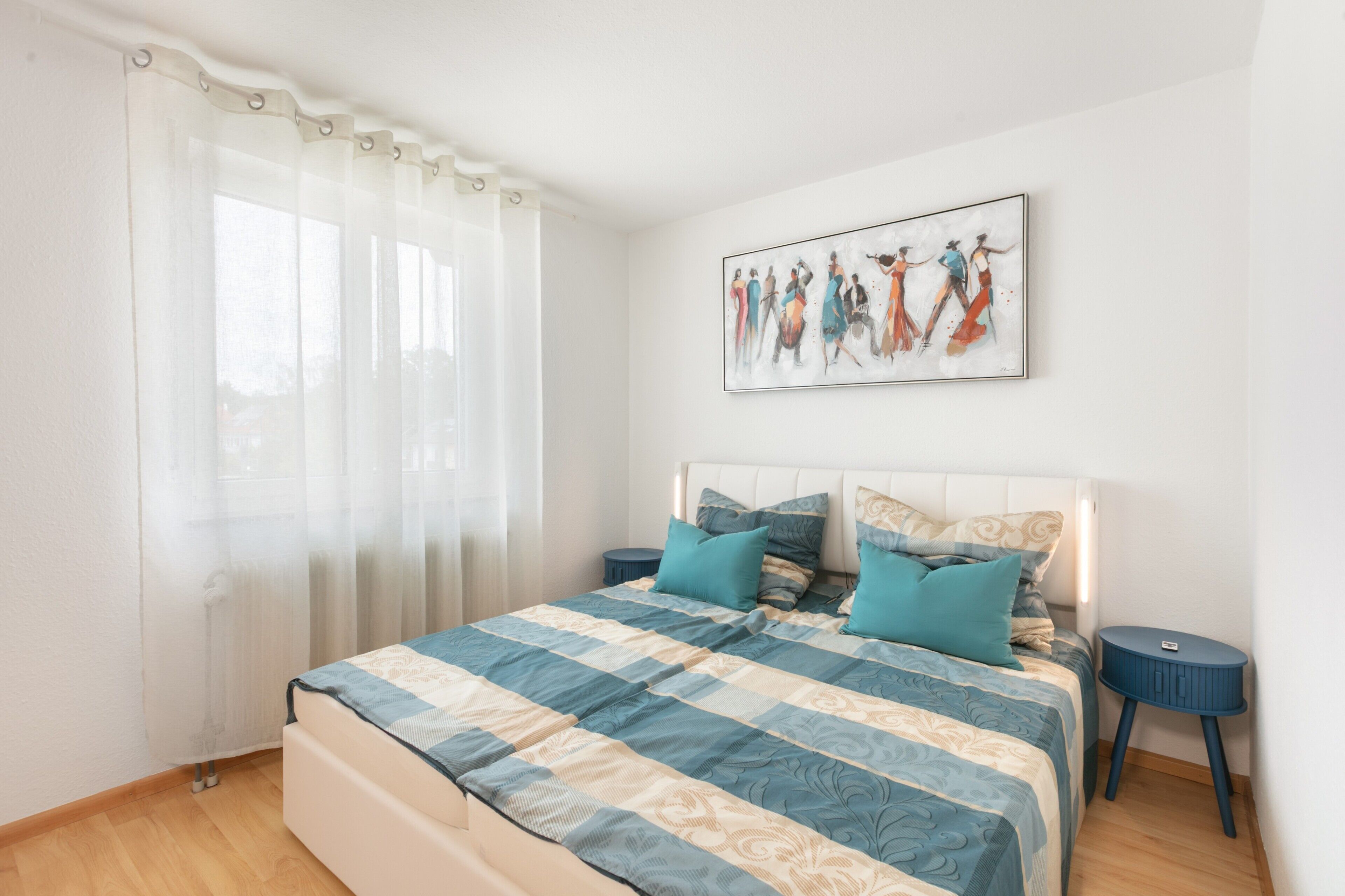Quarto com cama, almofadas, mesa de cabeceira, uma obra de arte na parede e janela com cortinas.