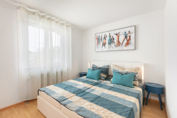 A bedroom with a bed, pillows, a nightstand, a wall art piece, and a window with curtains.