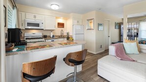 Une cuisine compacte avec un îlot central, deux tabourets de bar, un canapé blanc et un balcon avec vue.