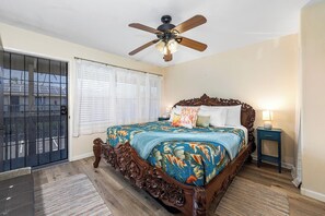 A bedroom with a large bed, a ceiling fan, a nightstand, and a view through a window with blinds. - Kona Shores#223: Top Floor, Split AC, Remodeled, Washer/Dryer (Kailua Kona)