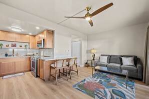 Eine moderne Küche mit Holzausstattung, einer zentralen Kochinsel mit Barhocker, einem Deckenventilator und einem grauen Sofa im Wohnbereich.