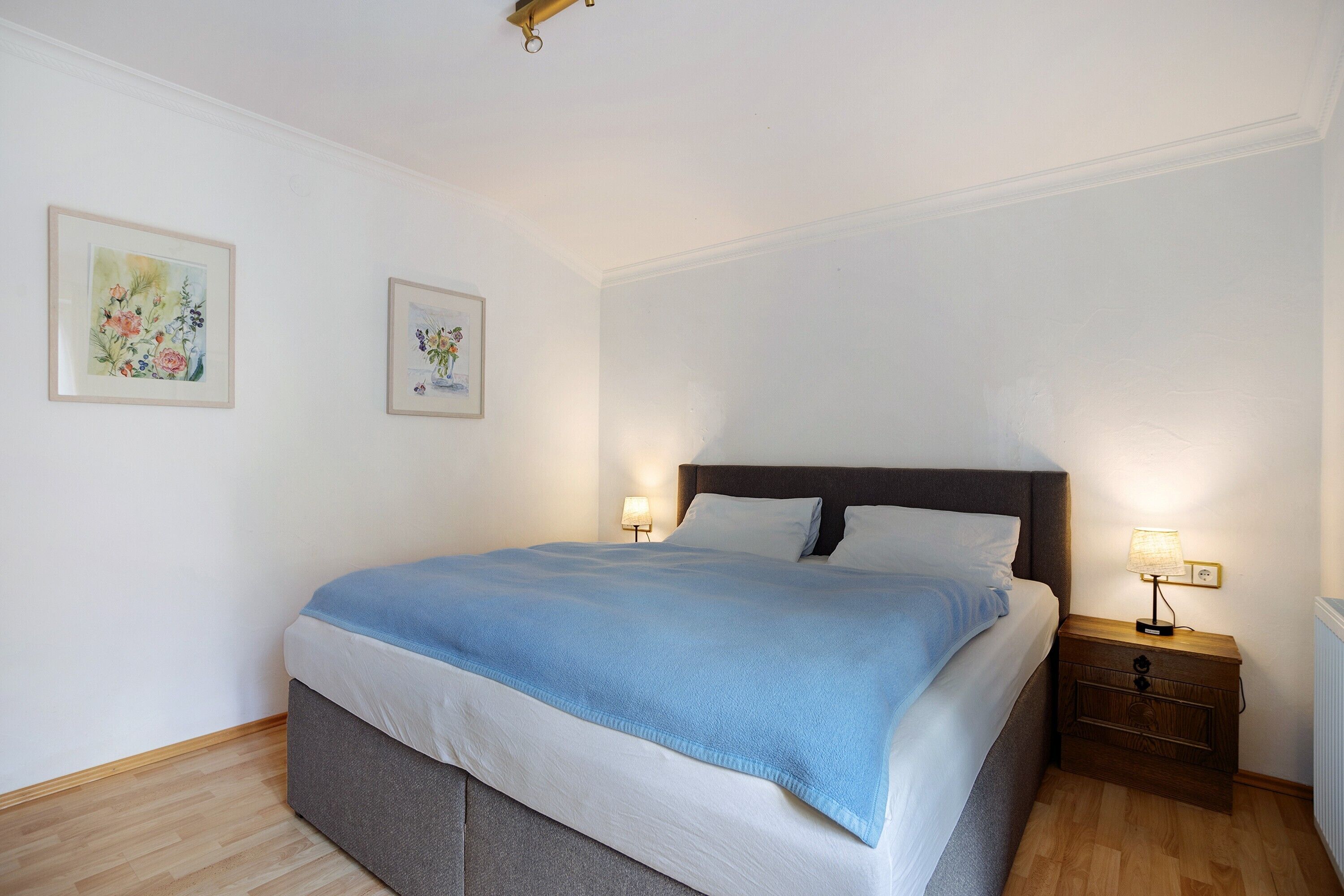 A neatly made bed with a blue blanket, two bedside lamps, and two framed floral paintings on the wall.