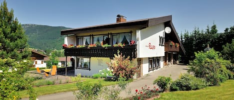 A traditional chalet-style building with a balcony, surrounded by a well-maintained garden and a mountainous backdrop.