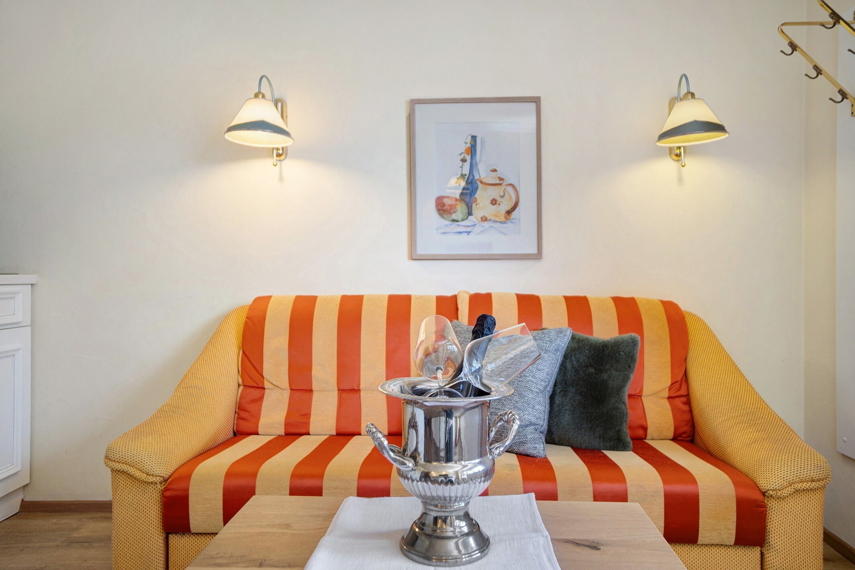 Una habitación con un sofá a rayas, dos lámparas fijadas a la pared, un cuadro enmarcado y una mesa con una jofaina de plata.