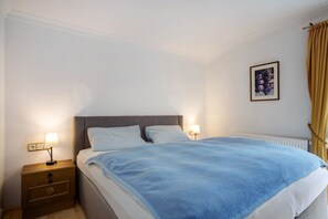 Un dormitorio con cama, lámparas de noche, mesita de noche, radiador y un cuadro en la pared.