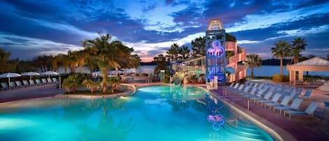 A resort pool area with lounge chairs, palm trees, and a water slide.