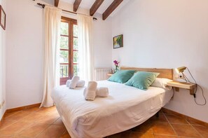 Un dormitorio con cama, almohadas, una mesita de noche con lámpara, ventana con cortinas y un radiador.