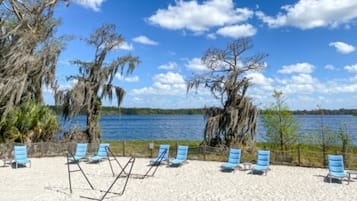 Een strand met blauwe stoelen en een zandvlakte, met uitzicht op een wateroppervlak met bomen en een heldere lucht.