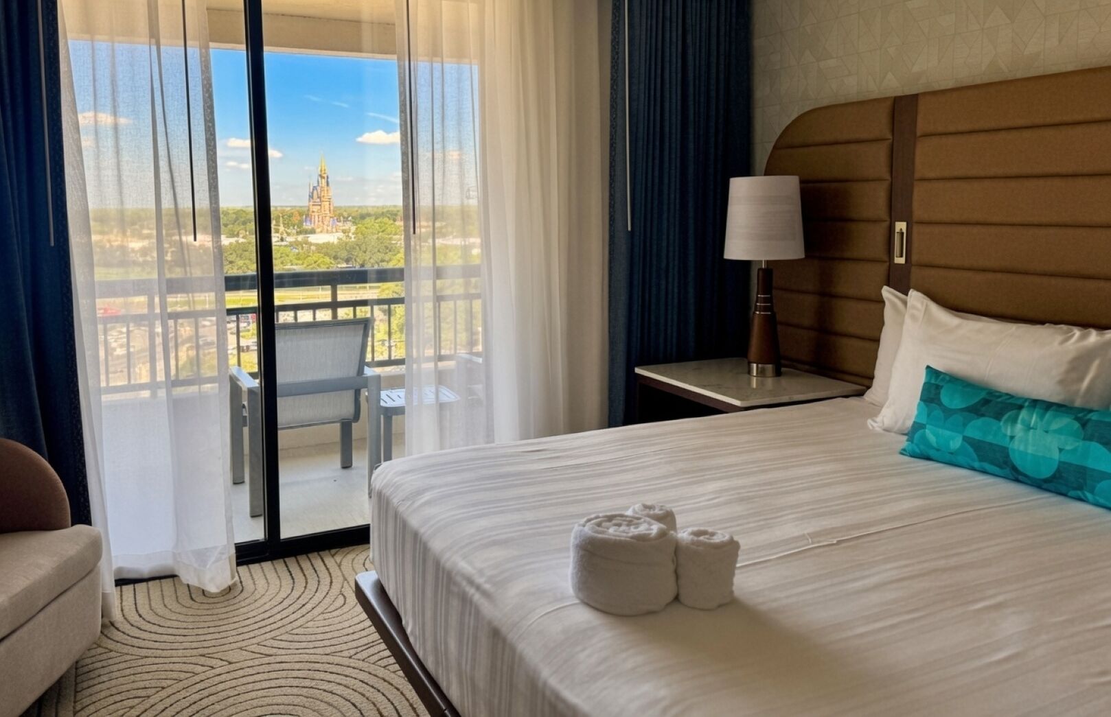 A hotel room with a bed, a nightstand, a lamp, a chair, and a view of a building through a window.