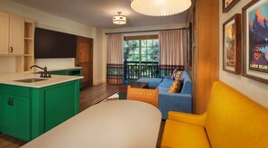 Ein kompakter Wohnbereich mit Küchenzeile, einem Sofa, einem gelben Sessel und einem großen Fenster mit Vorhängen.