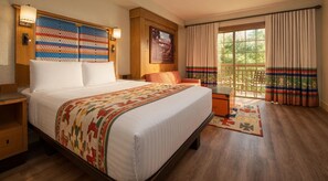 Ein Hotelzimmer mit einem großen Bett, einem Holfnachtkästchen, einem kleinen Tisch, einem Sessel und Blick ins Freie durch ein Fenster mit Vorhängen.