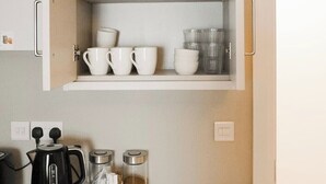 A kitchen with a white cabinet containing glassware, white dishes, and a kettle on the counter.
