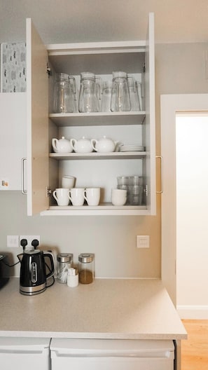 A kitchen with a white cabinet containing glassware, white dishes, and a kettle on the counter.