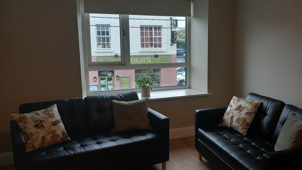 A room with two black leather sofas, a window with a view of a street and shops, and a radiator on the wall.