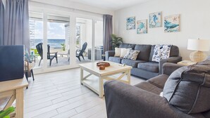 Ett vardagsrum med en soffa, ett soffbord och en tv. Det finns två fåtöljer och ett litet bord med en korg. Från rummet har man utsikt över havet genom stora glasdörrar.