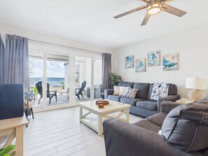 A living room with a sofa, a coffee table, and a TV. There are two chairs and a small table with a basket. The room has a view of the ocean through large glass doors. - SoulShine is oceanfront with beautiful sunrises and sunsets in St. Croix! (Christiansted)