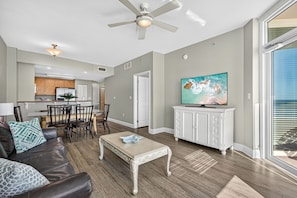 A modern living room with a dining area, a ceiling fan, a television on a cabinet, and a view through a large window. - Gulf-Front Condo with Balcony Access from the Primary Suite + Steps to Pier Park (Panama City Beach)