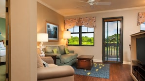 A living room with a sofa, a coffee table, a lamp, a TV stand, and a window with a view of greenery. - Hilton Head | Couples Island Retreat | Pool + Beach (Hilton Head Island)