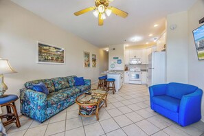 Ein Wohnzimmer mit einer Couch, einem Couchtisch und einem blauen Sessel. Es gibt eine Küchenzeile mit weißen Schränken und einem Kühlschrank.