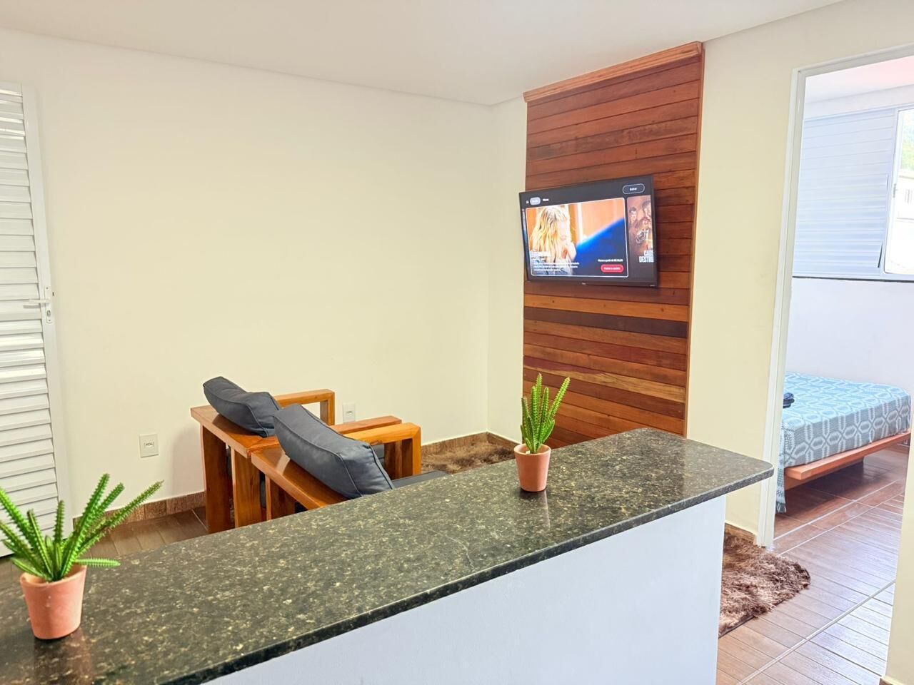 Un salon moderne avec un téléviseur à écran plat fixé à un mur en bois, un petit coin salon comprenant un canapé et une table basse, et un comptoir de cuisine agrémenté de plantes en pot.